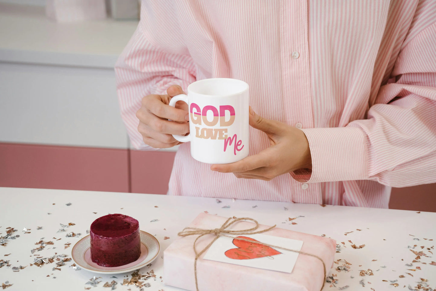Person holding a mug with 'GOD LOVE ME' text, sitting at a table with a cake and gift.
