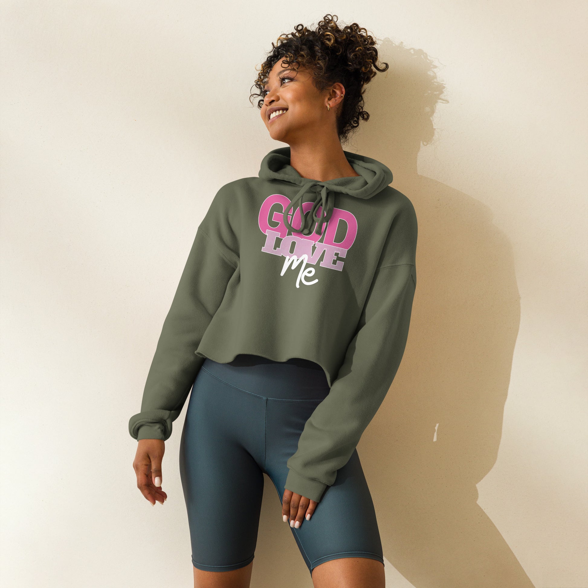 Woman wearing a green hoodie with pink text : God Love Me and dark shorts on a plain background
